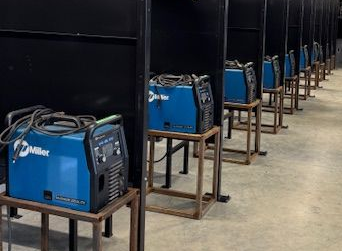 Welding shop with multiple blue welding machines on stands, work tables, and individual work stations.