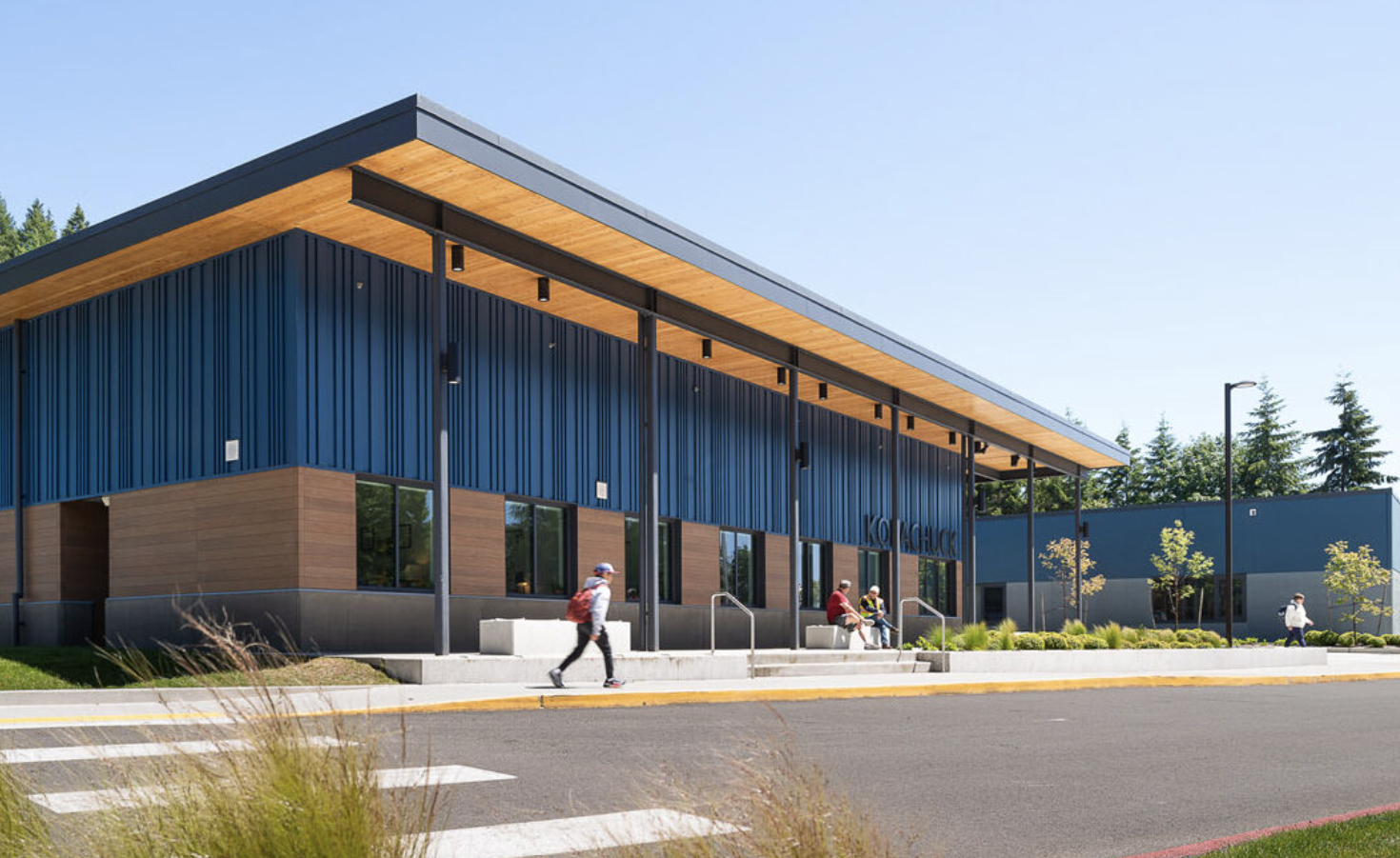 Modern building with blue siding, wood accents, and covered entrance; person walks toward the entrance.