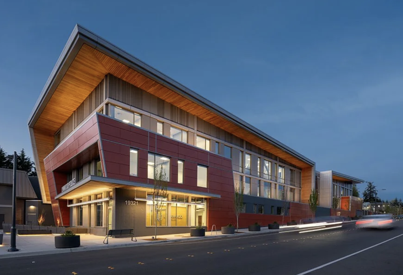 Lynwood Police station building, modern design, lit at dusk. Red, brown, and glass facade, flat roof.