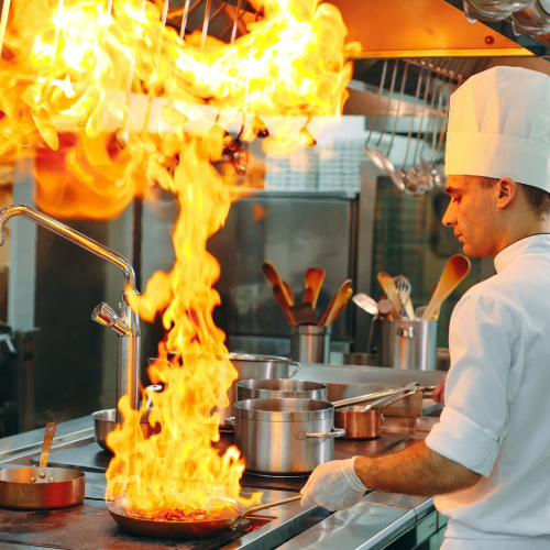 A chef near a stove cooking off alcohol that is on fire in a frying pan