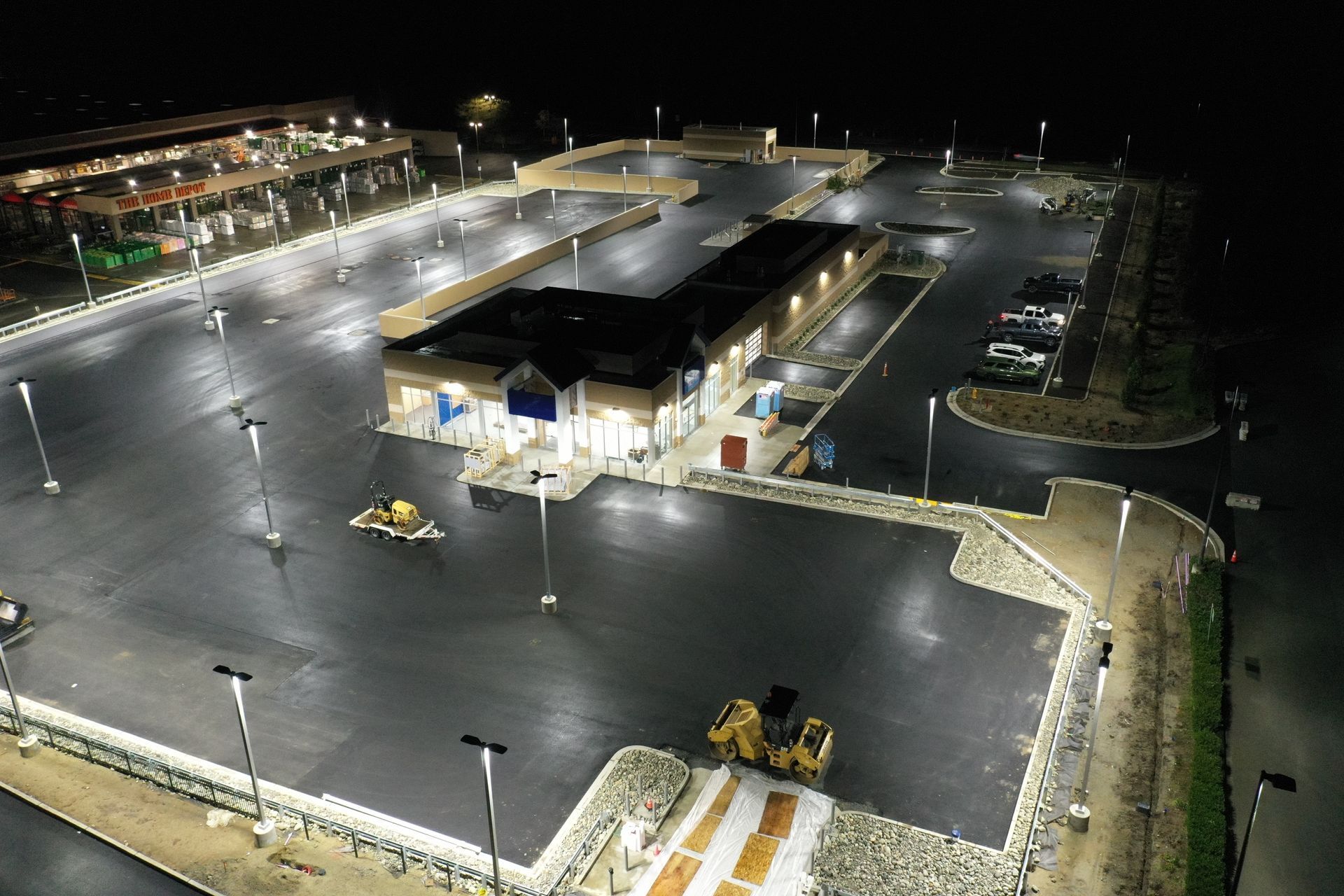 Night aerial view of a brightly lit commercial building with a large, empty asphalt parking lot.