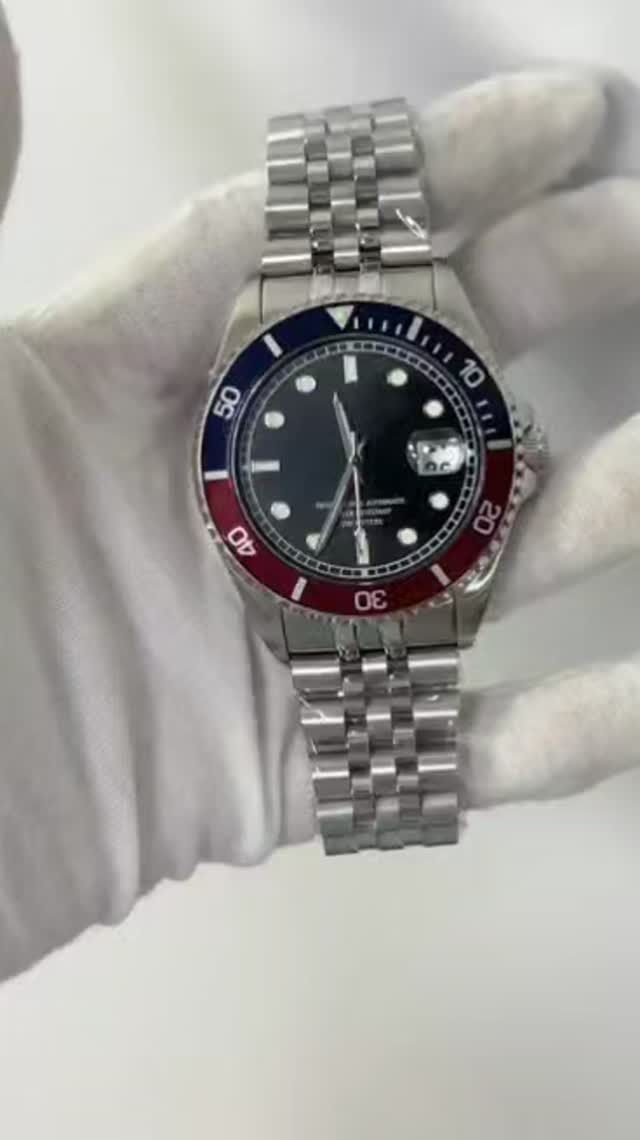 Reloj de pulsera plateado con esfera negra, bisel azul y rojo y pulsera de plata, sostenido por una mano enguantada.