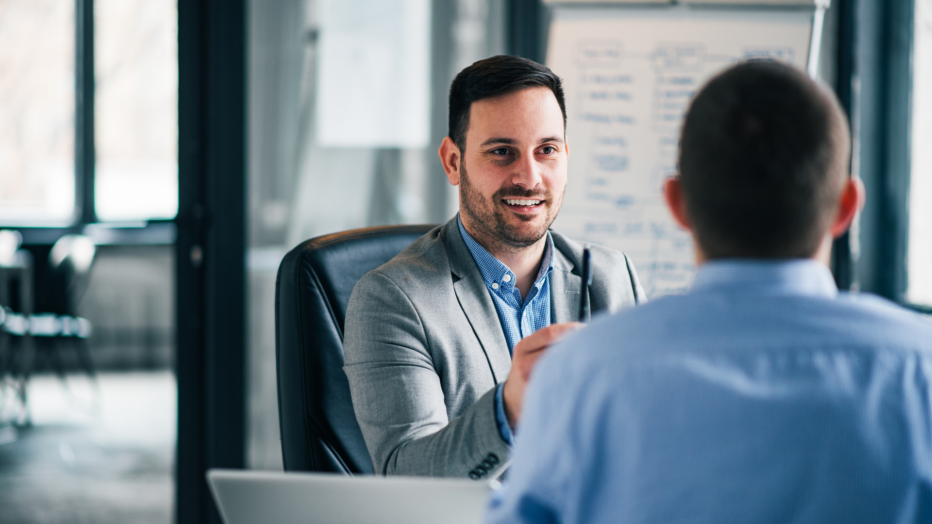 A man is sitting in a chair talking to another man in an office.