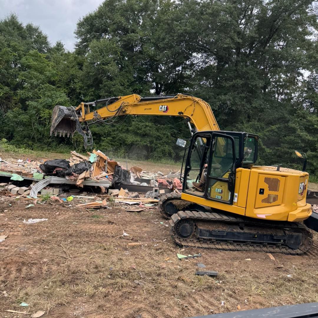 Yellow excavator demolishing a structure outdoors on a brown field; trees in the background.