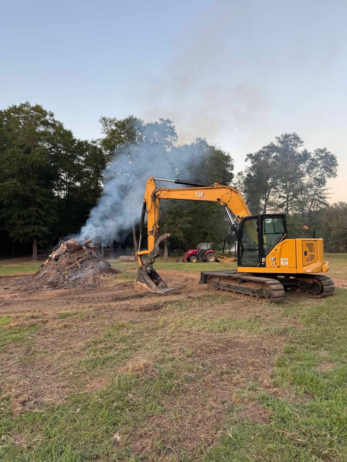 Skid steer compacting gravel on a dirt road, near trees and a wooden fence.