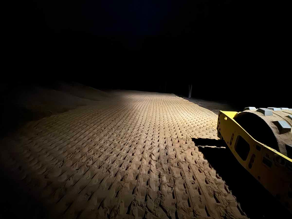 Night view of a yellow construction roller on a freshly laid road. Headlights illuminate the textured surface.