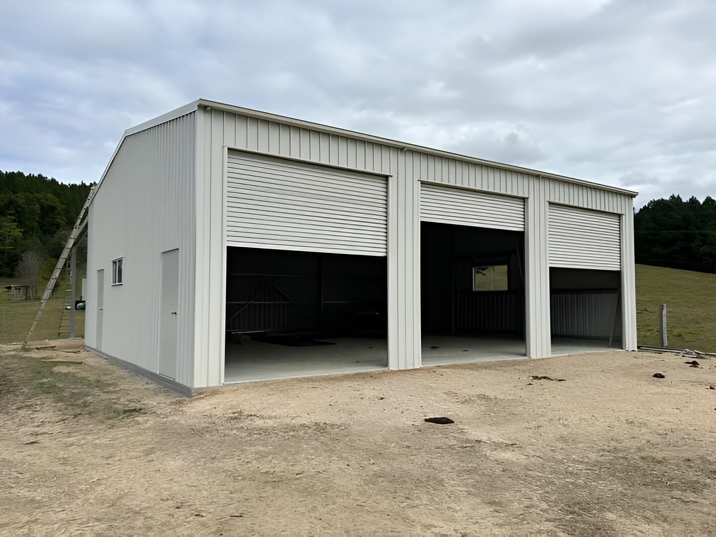 A Large White Building With Three Garage Doors Is Sitting On Top Of A Dirt Field — Mbah Metal Roofing In Murwillumbah, NSW