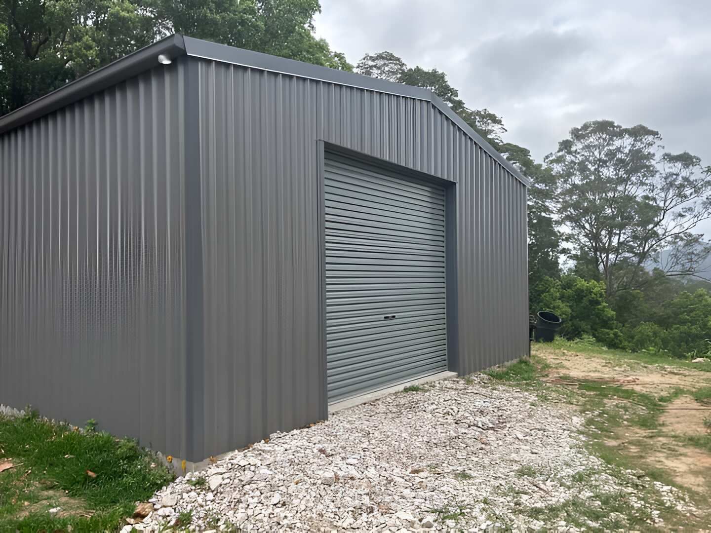 A Gray Metal Building With A Garage Door Is Sitting On Top Of A Gravel Road — Mbah Metal Roofing In Murwillumbah, NSW