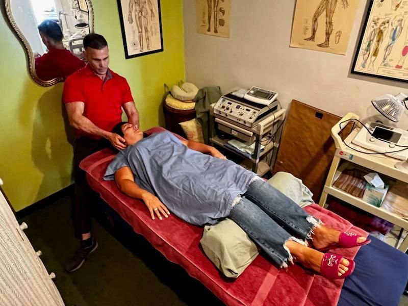 A practitioner in a red shirt performs a physical adjustment on a person lying on a red massage table in a clinic.
