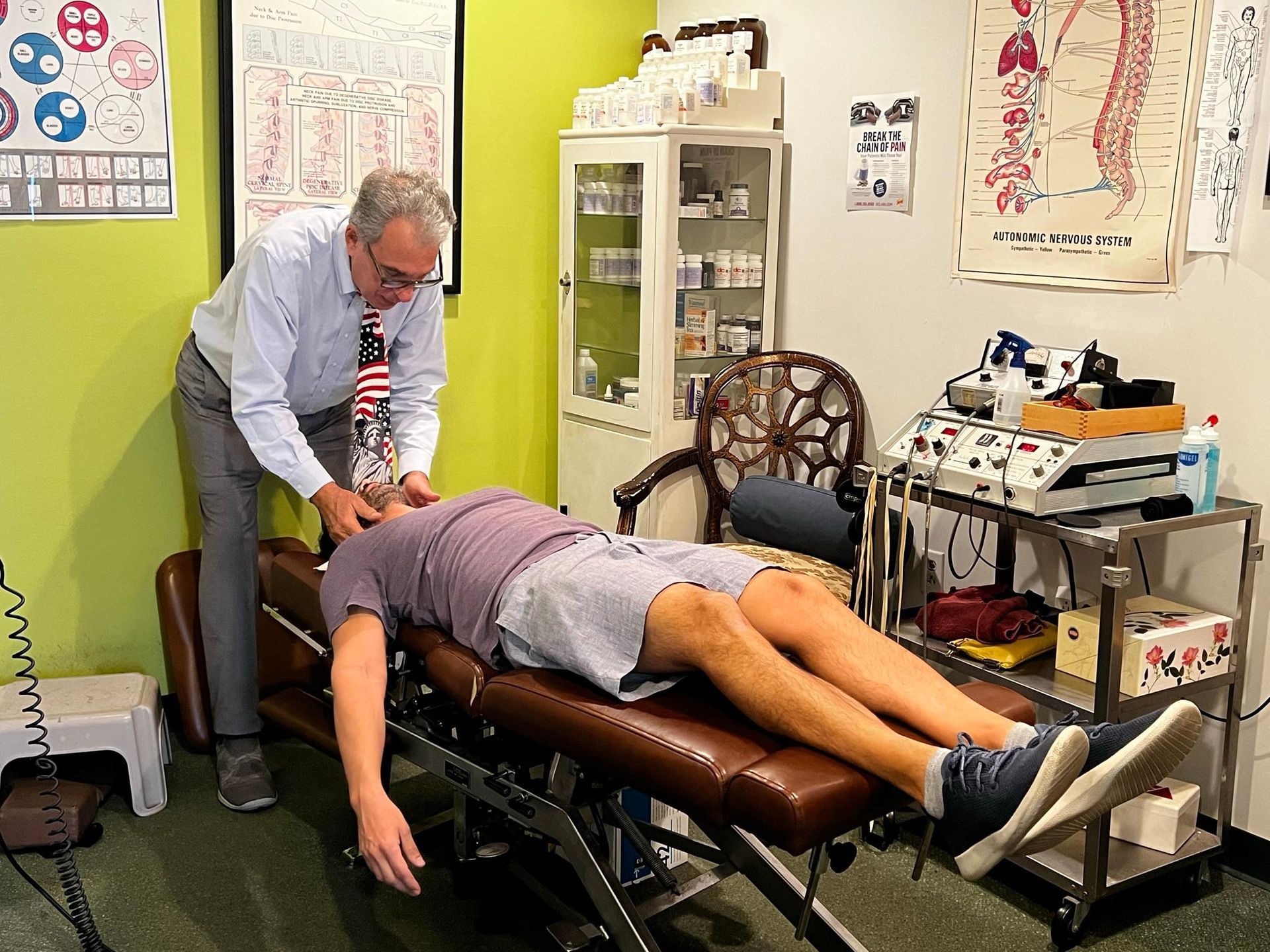 A professional adjusts a patient lying face down on a clinical table in an office setting with medical charts on walls.