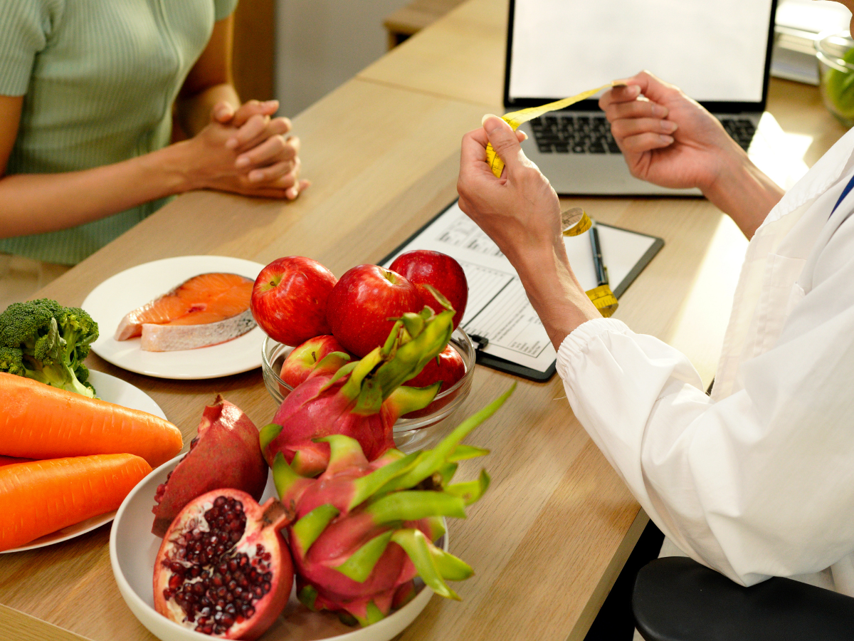 A professional consults with a patient at a desk featuring bowls of fresh fruit, vegetables, and fish for dietary planning.