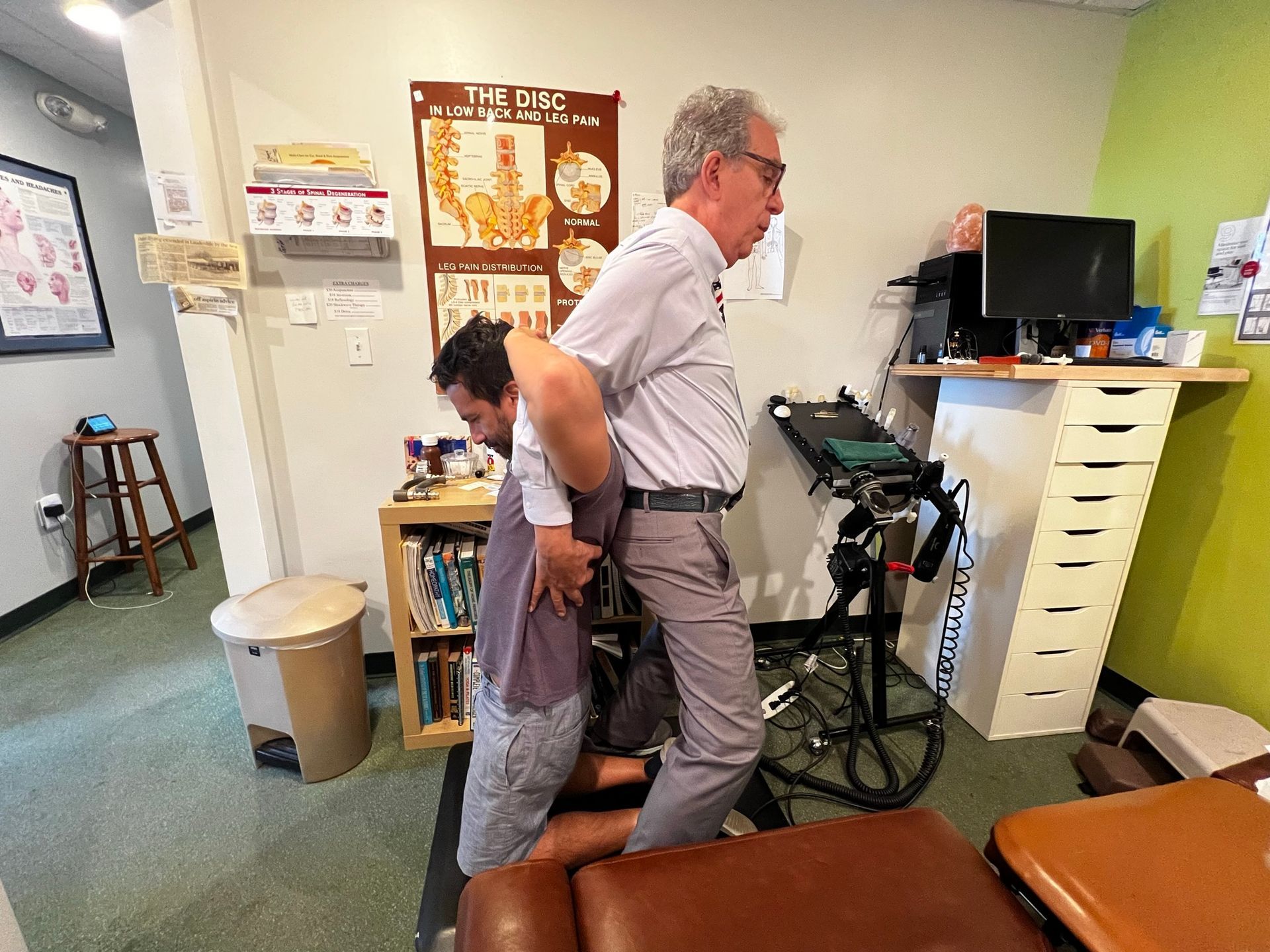 A chiropractor performs a spinal adjustment on a person kneeling on a table in an office setting.