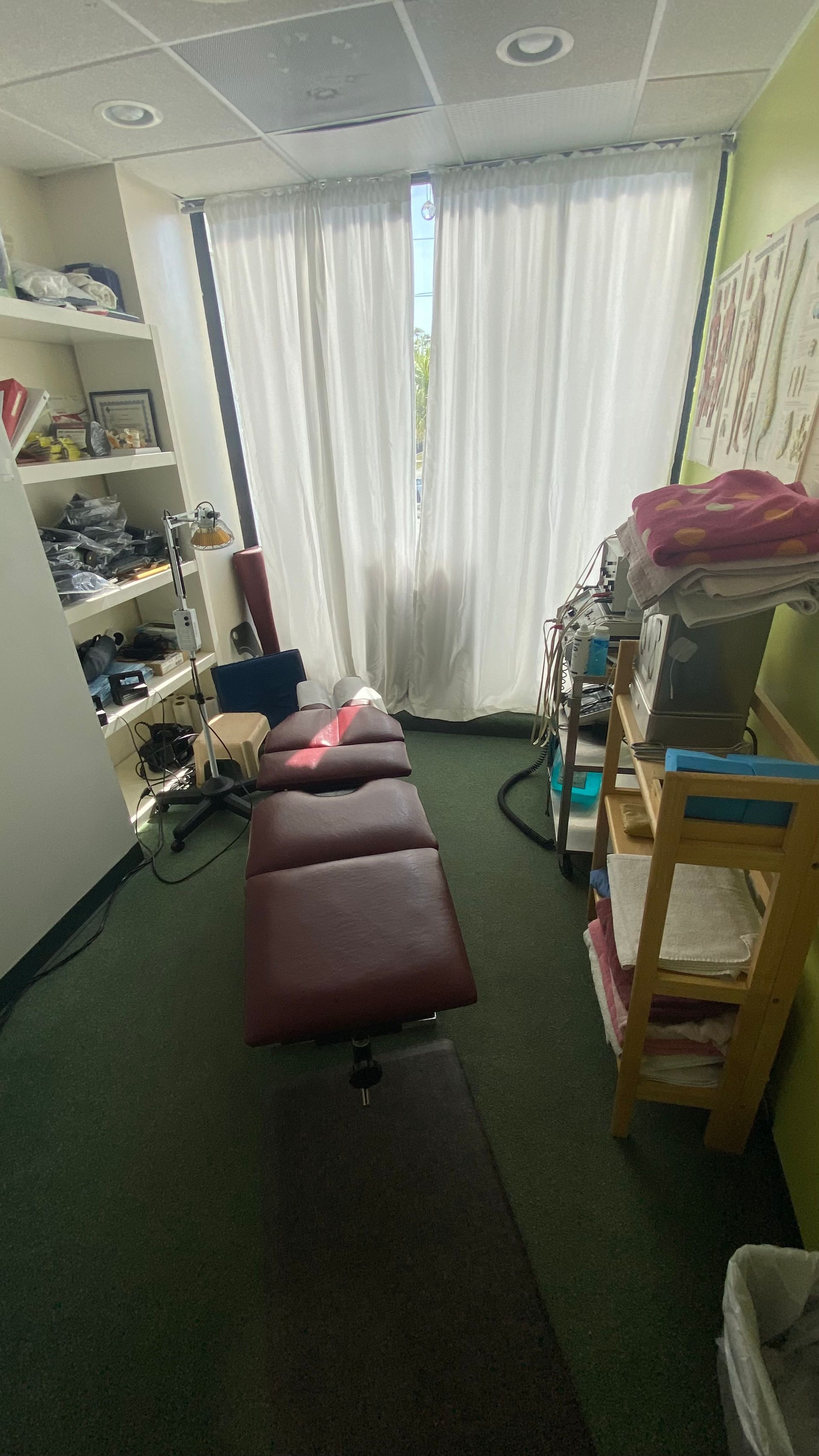 A treatment room features a burgundy massage table centered in the space, flanked by shelves and a cart against the walls.
