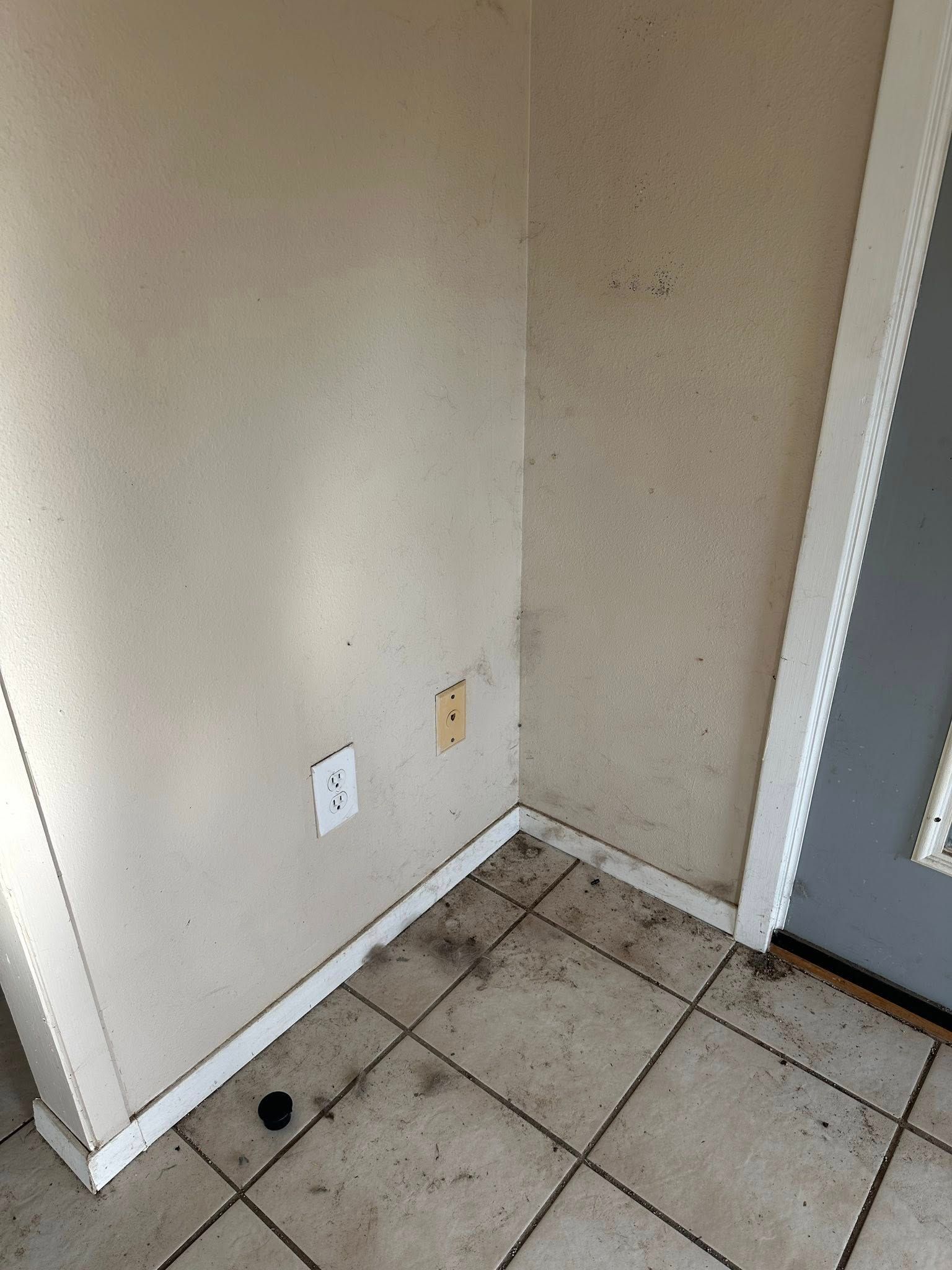 Corner of a room with dirty tile floor, off-white walls, and a door frame. Electrical outlets visible.