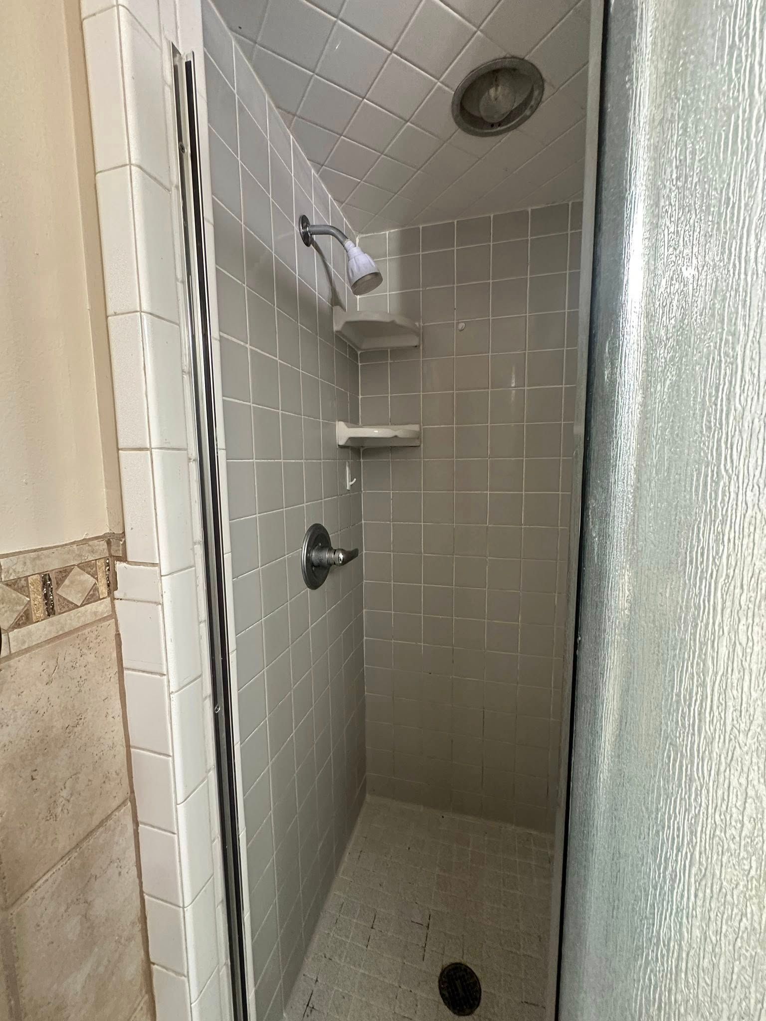 Small shower with gray tiled walls, showerhead, and corner shelves.
