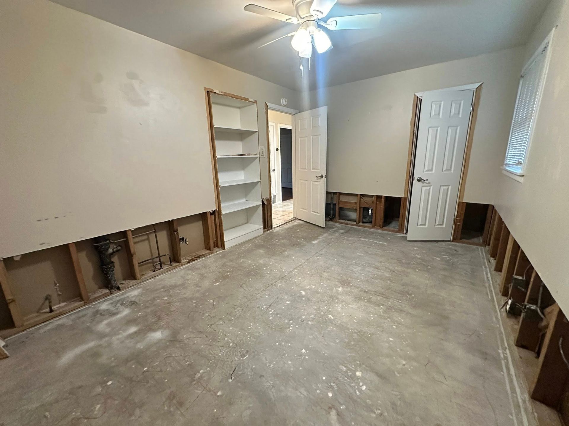 Room with exposed wall studs, concrete floor, and partially built-in shelving. Drywall removed from lower walls.