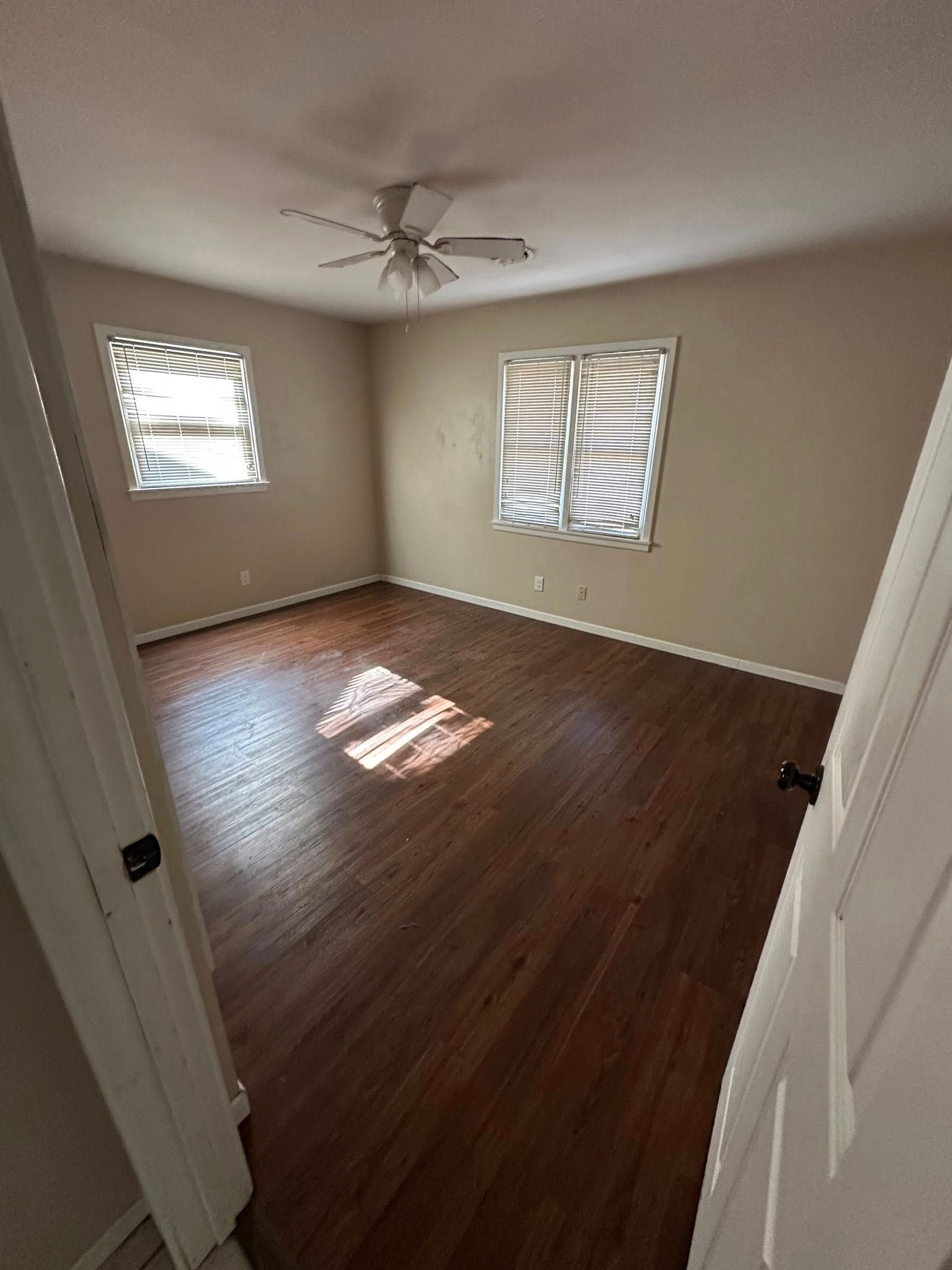 Empty bedroom with brown hardwood floors, two windows with blinds, and a ceiling fan.
