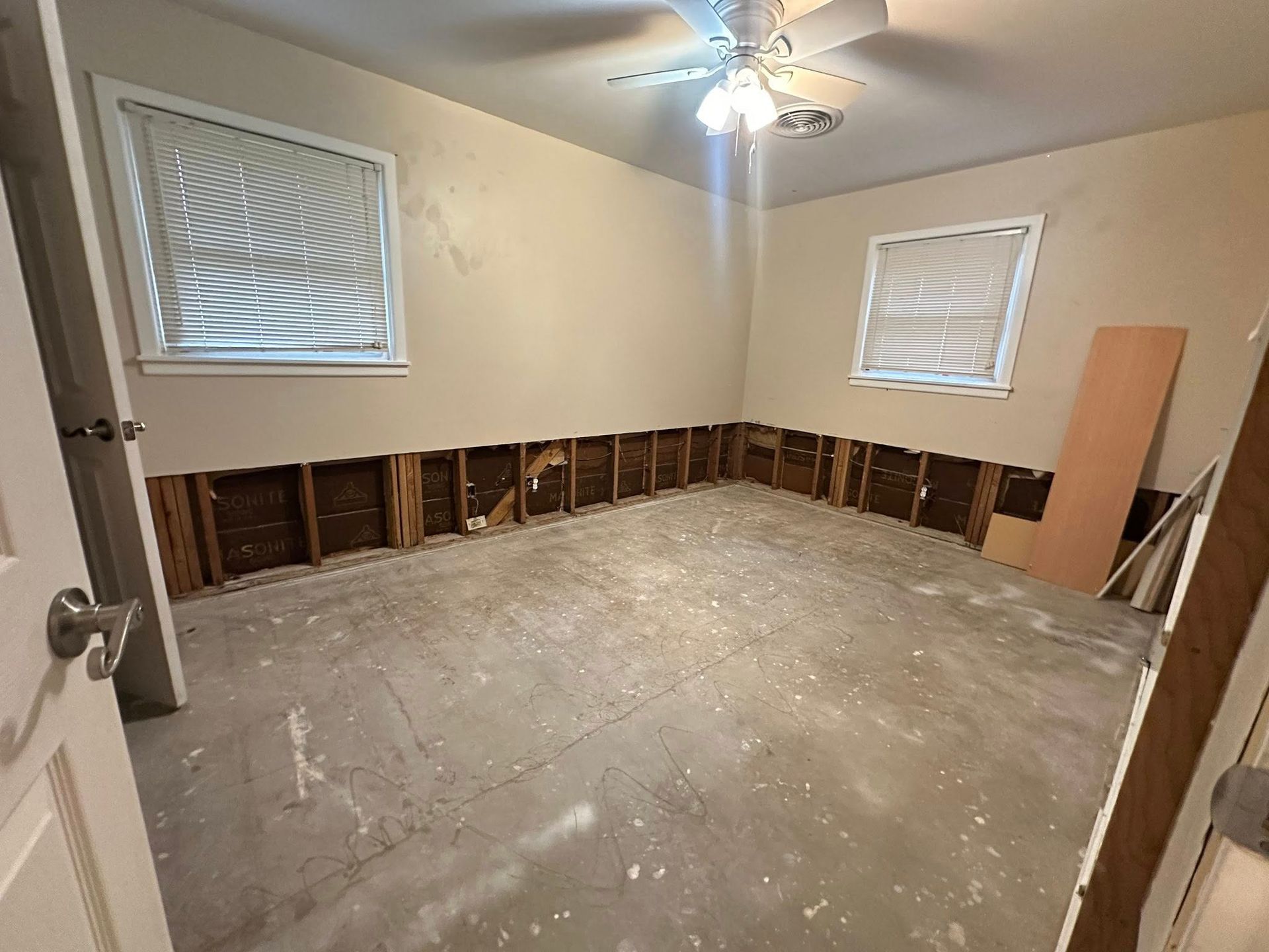 Empty room undergoing renovation with exposed studs, two windows, and debris on the floor.