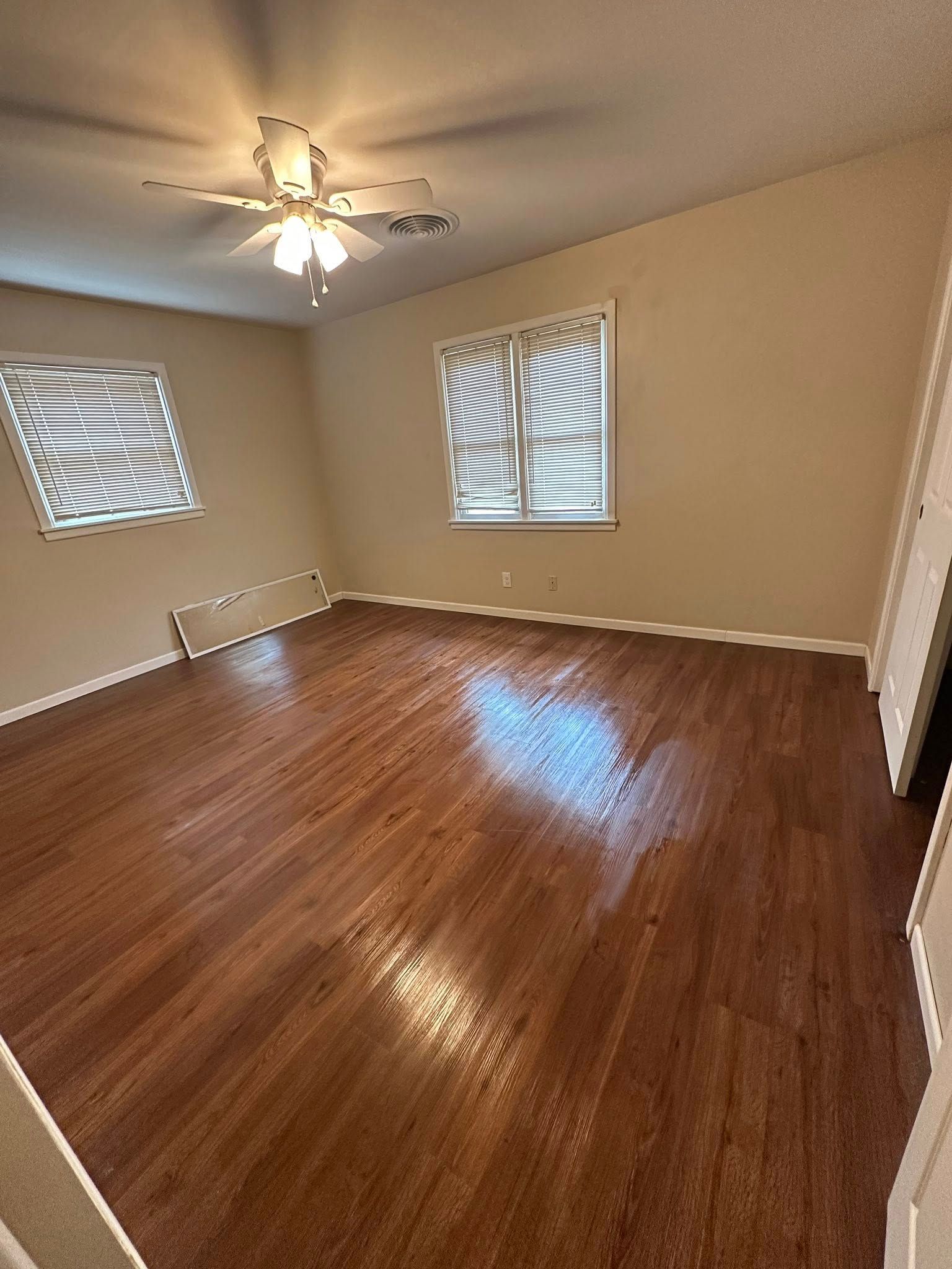 Empty room with hardwood floors, two windows with blinds, and a ceiling fan.