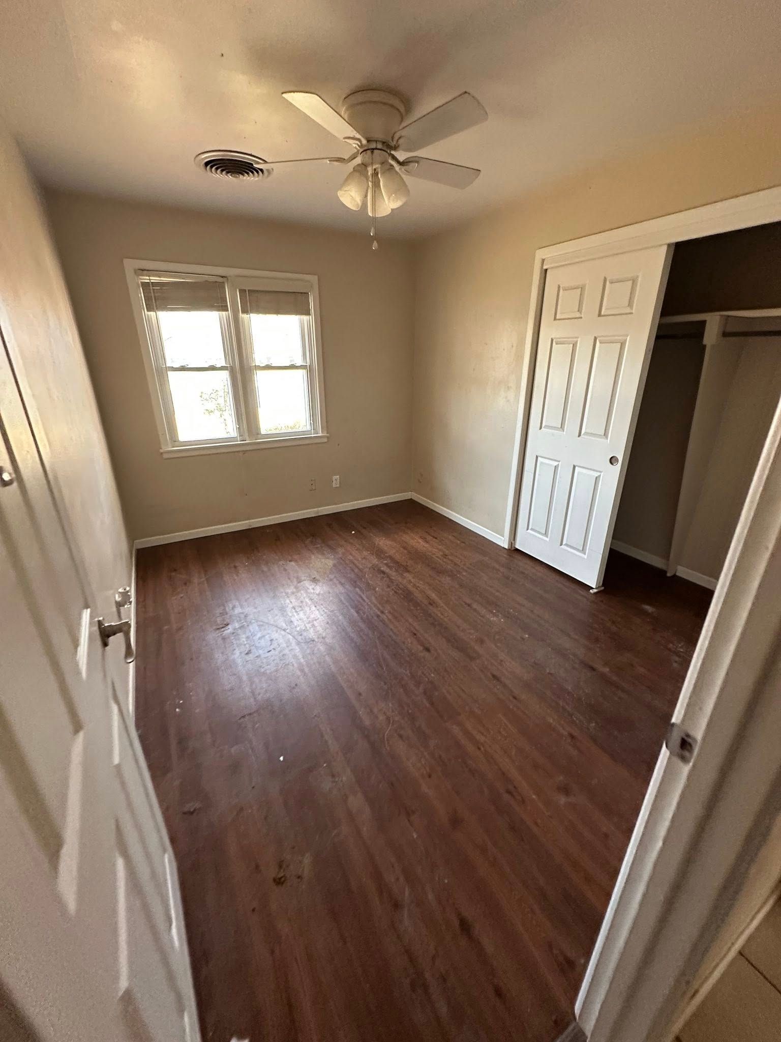 Empty bedroom with wooden floor, white ceiling fan, closet, and window. Beige walls.