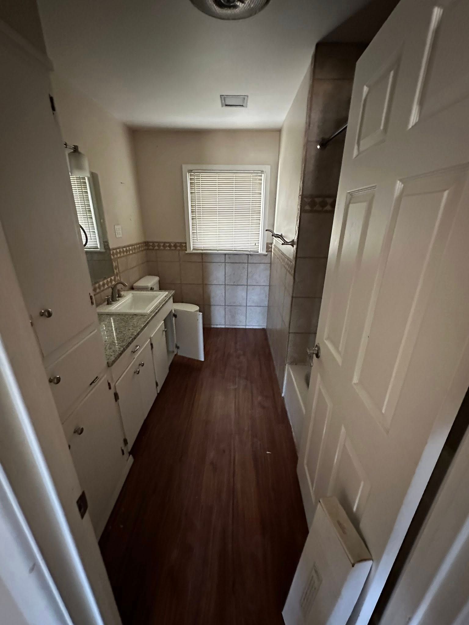 Bathroom with white cabinets, dark wood floor, and a window with blinds.