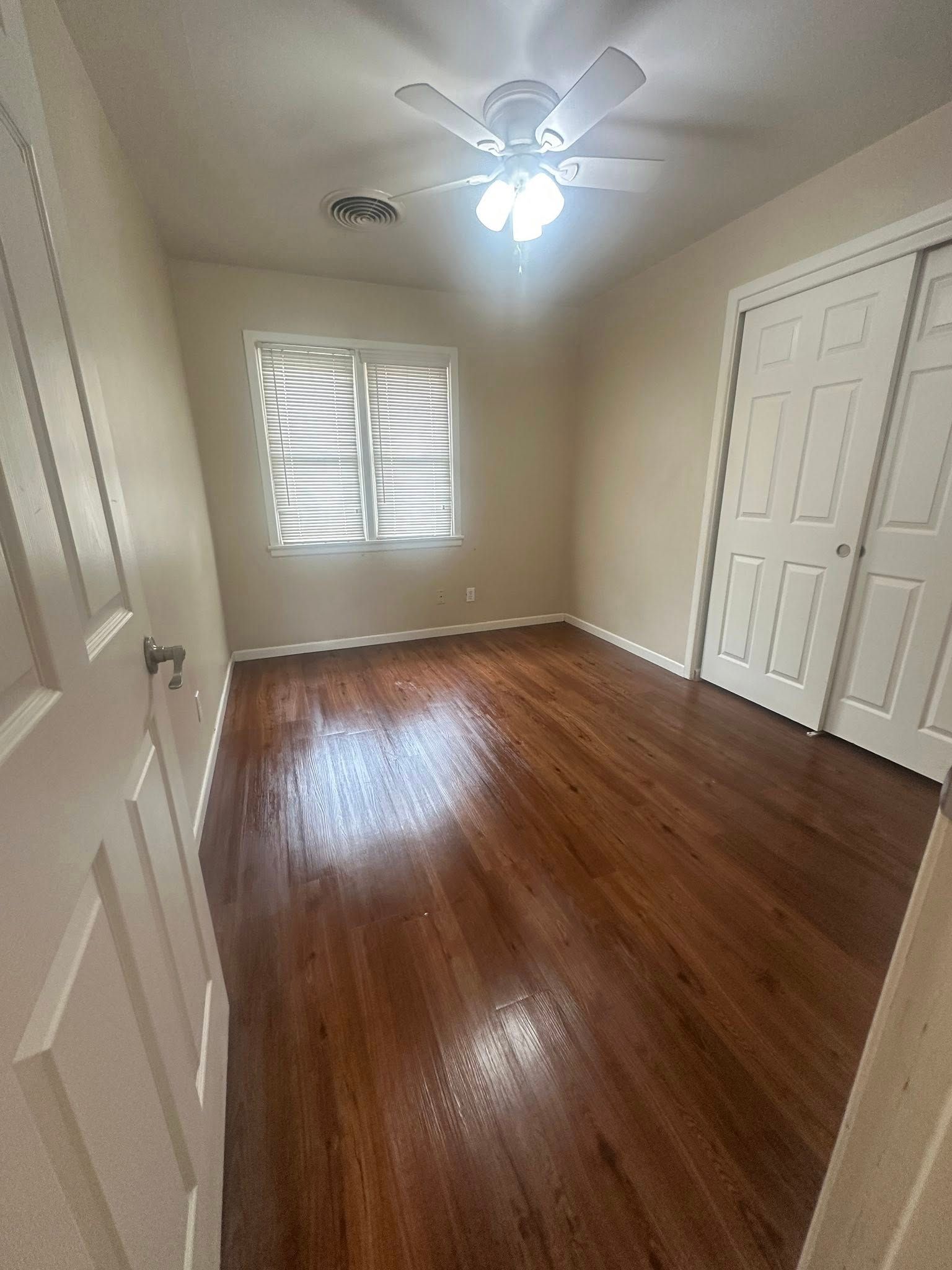 Empty bedroom with hardwood floors, white sliding closet doors, and a window with blinds.