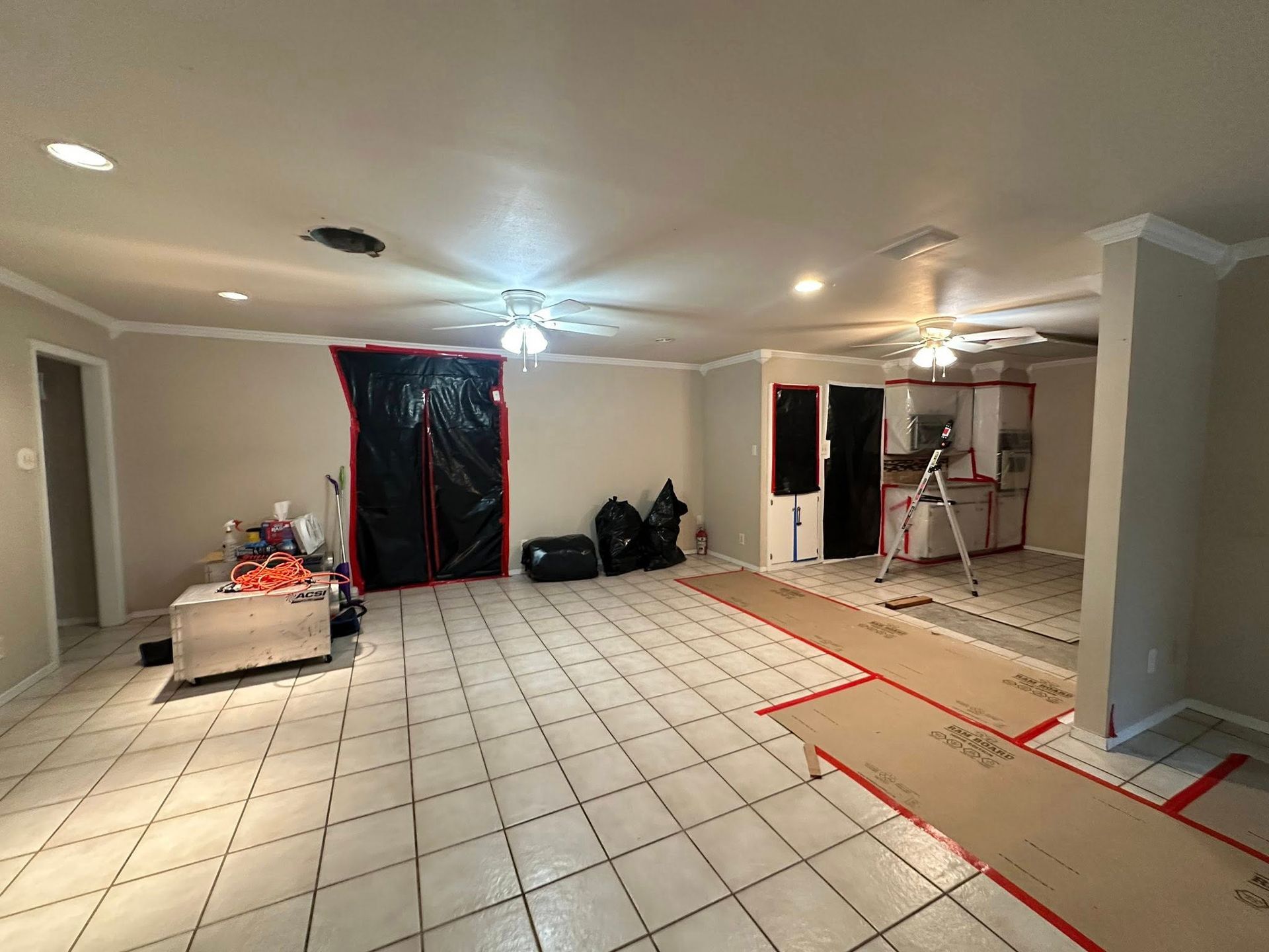 Room renovation in progress, with protective plastic sheeting, cardboard, and tools on tile floor.