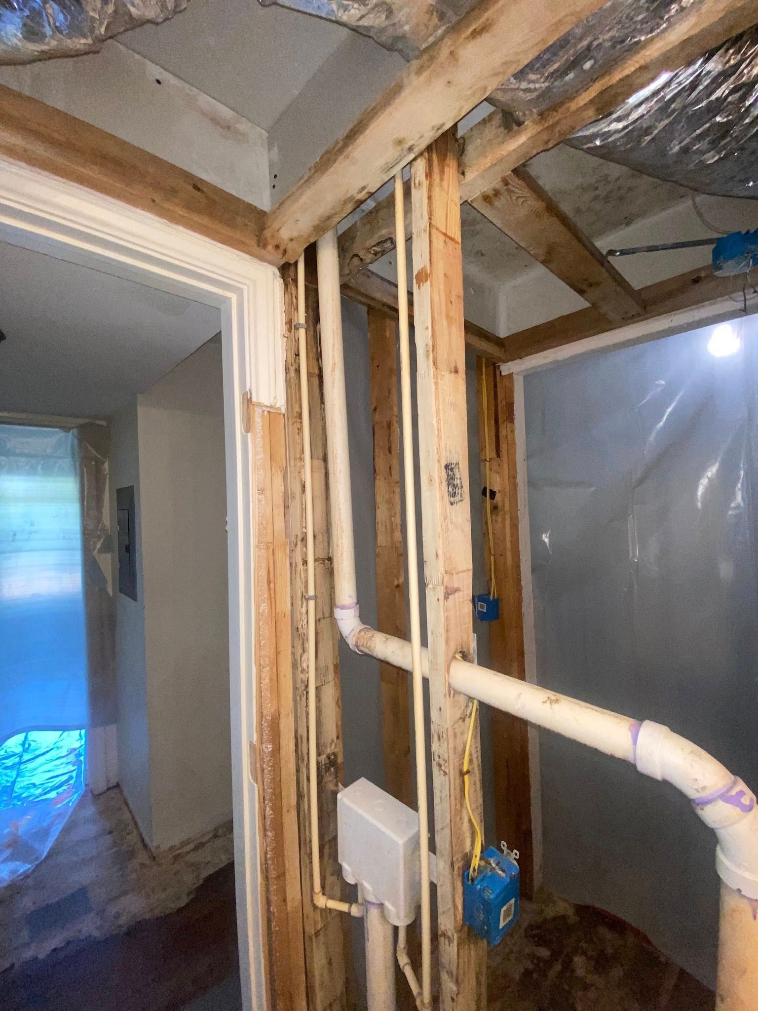 Interior view of a bathroom remodel in progress, showing exposed framing, plumbing, and electrical work.
