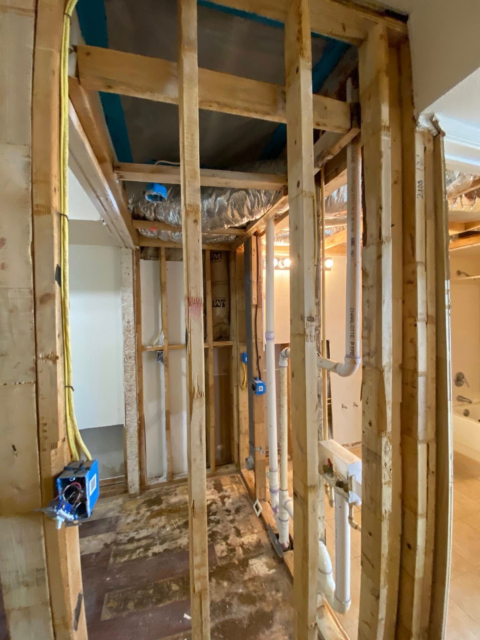 Interior view of a bathroom under construction, showing wooden framing, exposed pipes, and wiring.