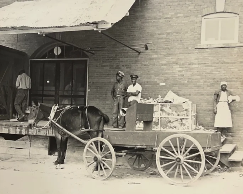A horse-drawn wooden cart loaded with cargo stands beside a brick building, with several figures nearby.