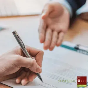 Person signing a document with another person's hand extended in the background. Close up.