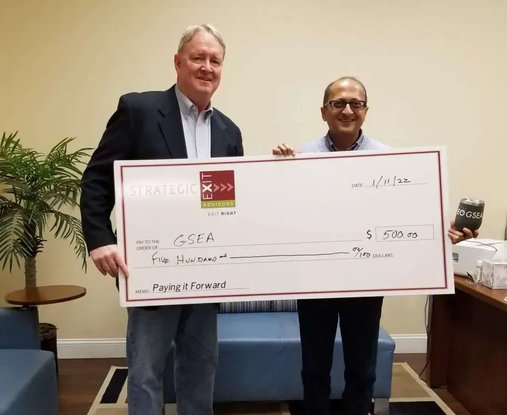 Two men holding an oversized check. One man in a suit, the other in a button-up shirt. Indoor office setting.