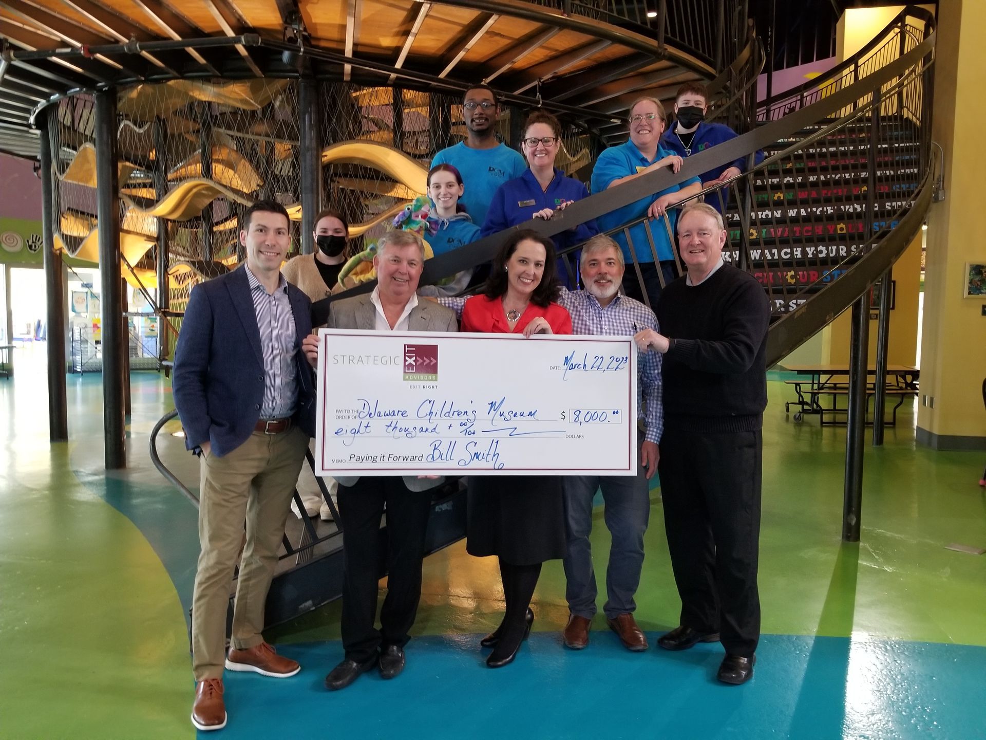 Group of people holding a large check in a colorful building with a spiral staircase.