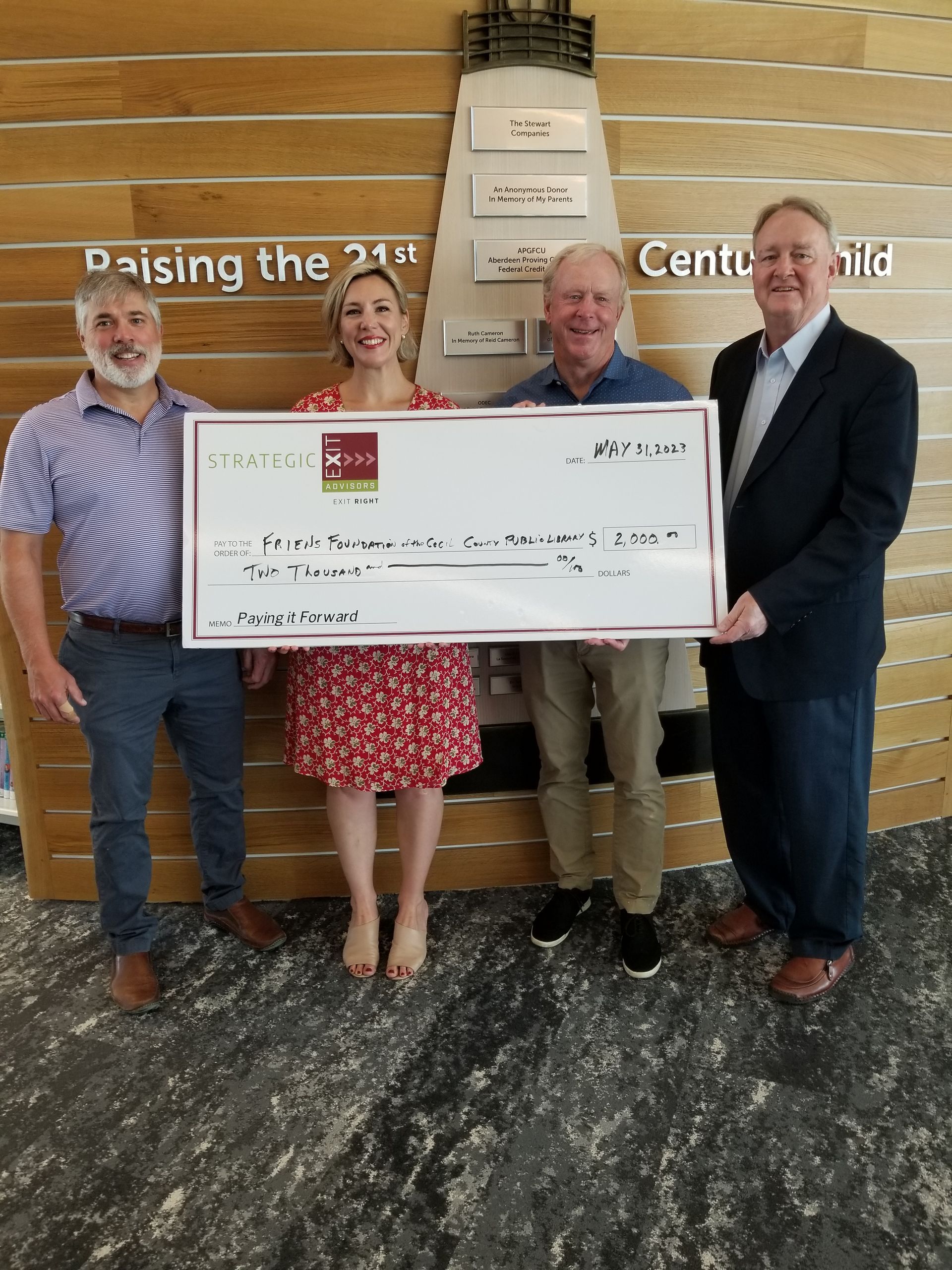 Four people holding a large check indoors, smiling. They stand in front of a wooden wall with raised lettering and art.
