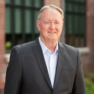 Man in a suit smiles outdoors, a brick building in the background.