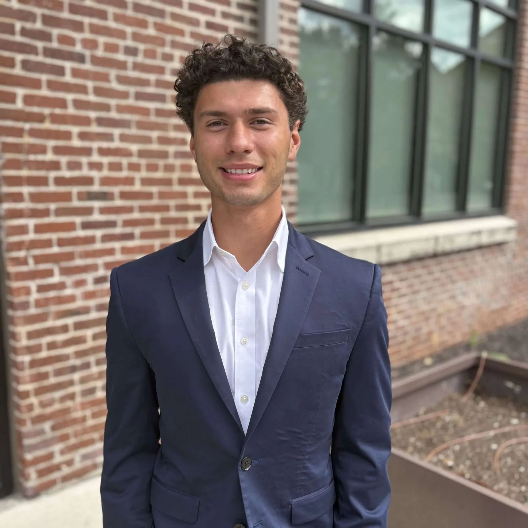 Man in a navy blazer smiles, standing in front of a brick building and large window.
