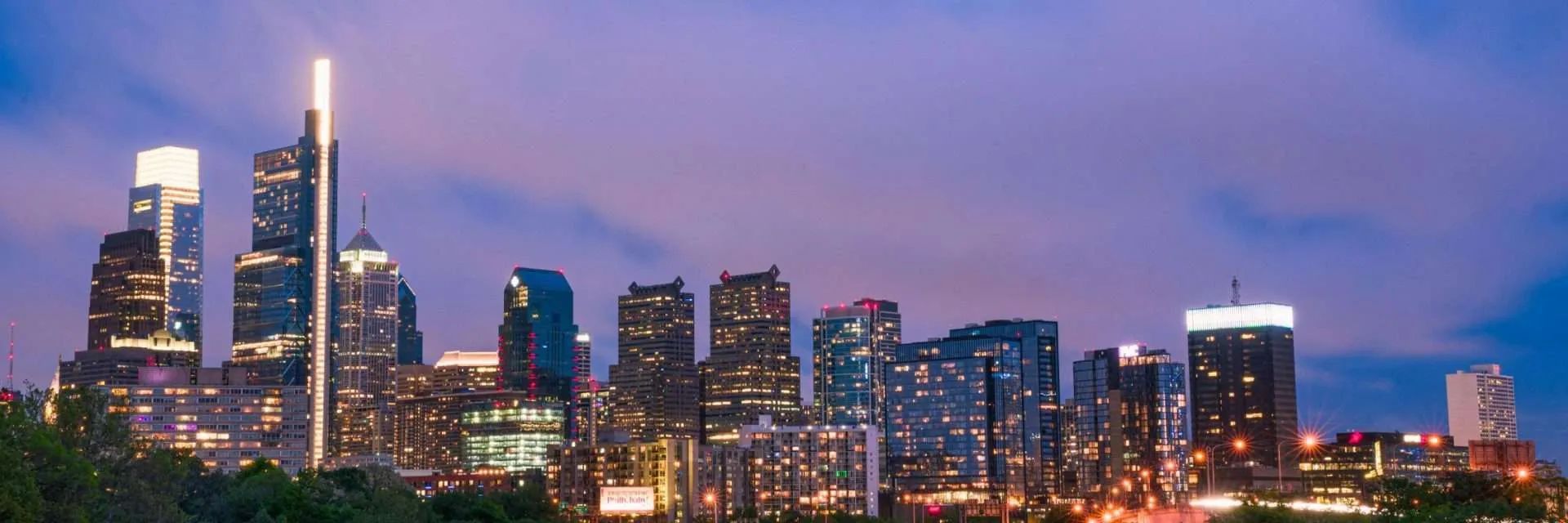 Philadelphia skyline at dusk, lit buildings under a purple sky.