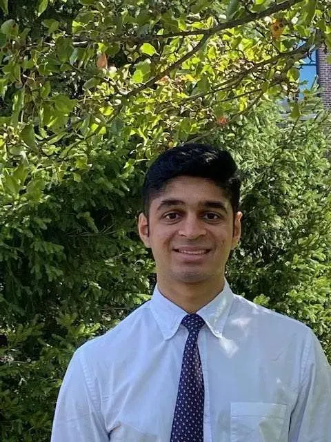 Man in a white shirt and patterned tie smiling in front of green foliage.
