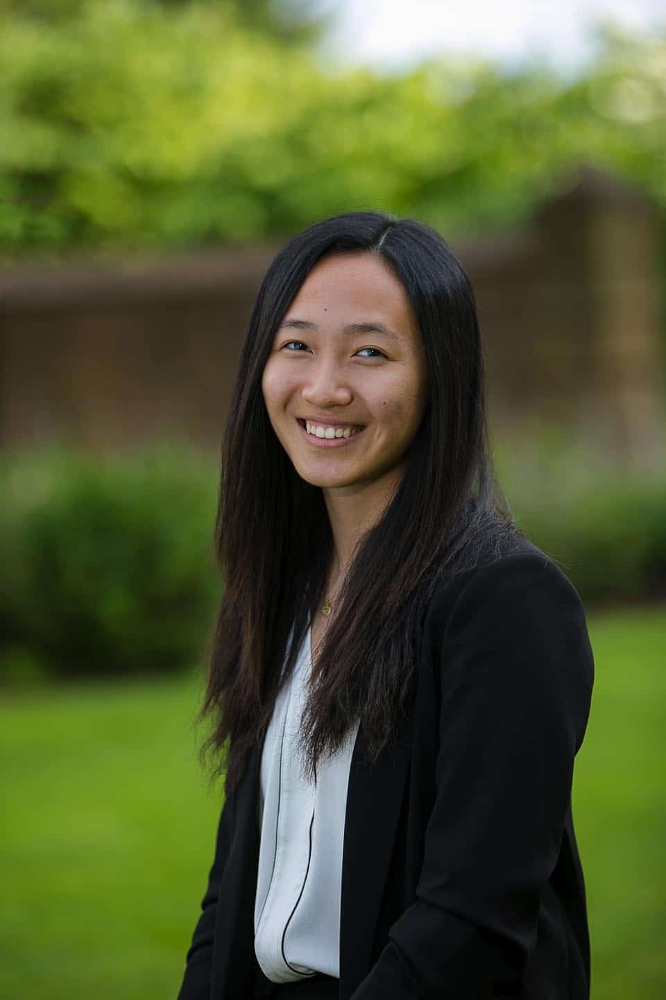 Woman in black blazer smiles outdoors with green background.