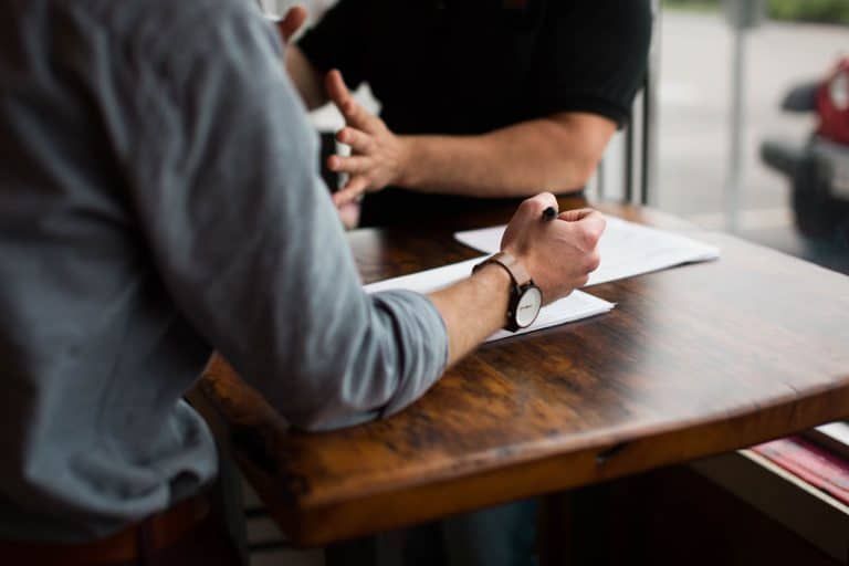 Two people at a table, one writing on a paper, the other gesturing.