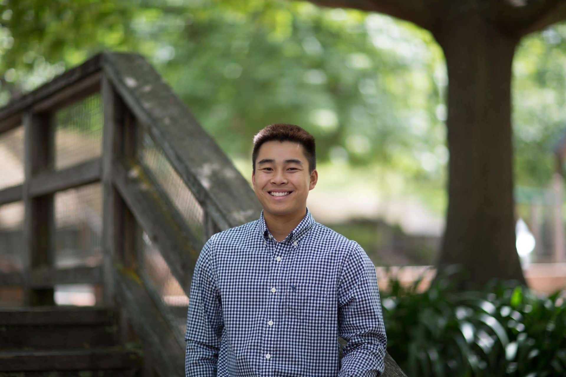 Man in blue plaid shirt smiles near wooden stairs and trees.