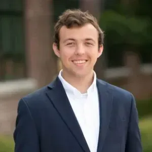 Smiling man in a navy suit jacket and white shirt, standing outside a brick building.