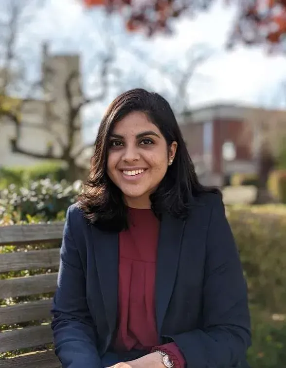 Woman in a navy blazer and maroon shirt smiles outdoors, seated on a wooden bench, with a building in the background.