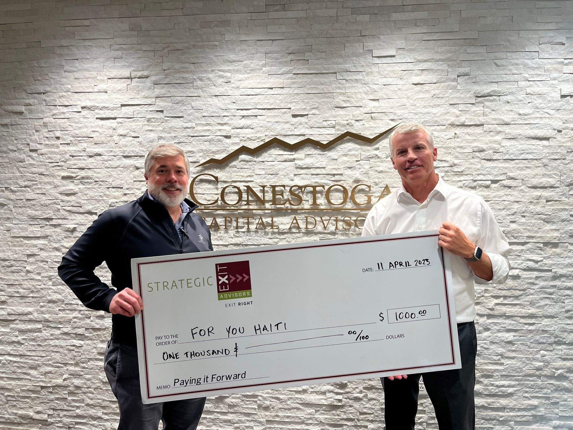 Two men holding a large check with the logo of a company. They are standing in front of a wall with the company name on it.