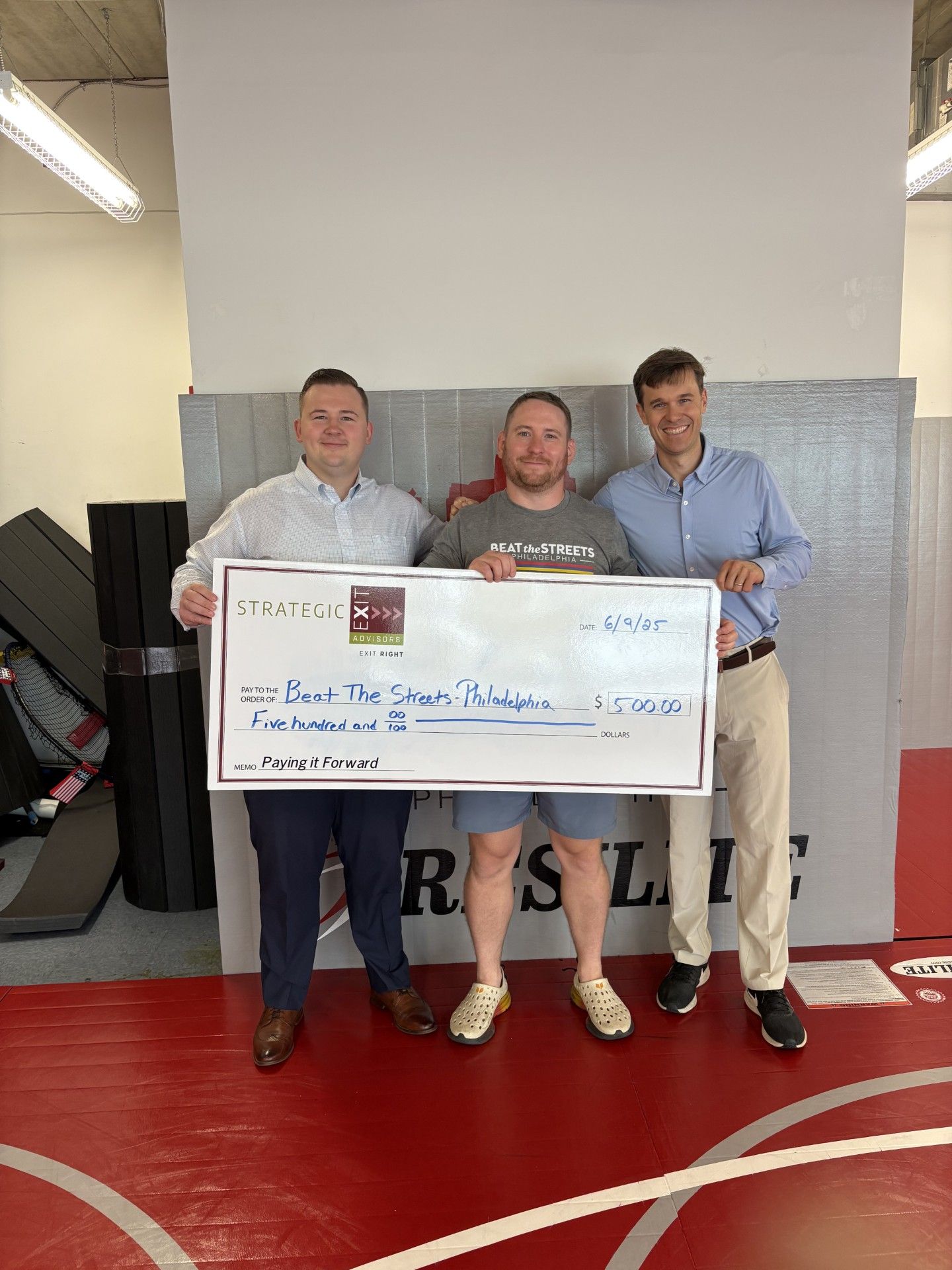 Three men holding a large check at a wrestling gym. The check is for 