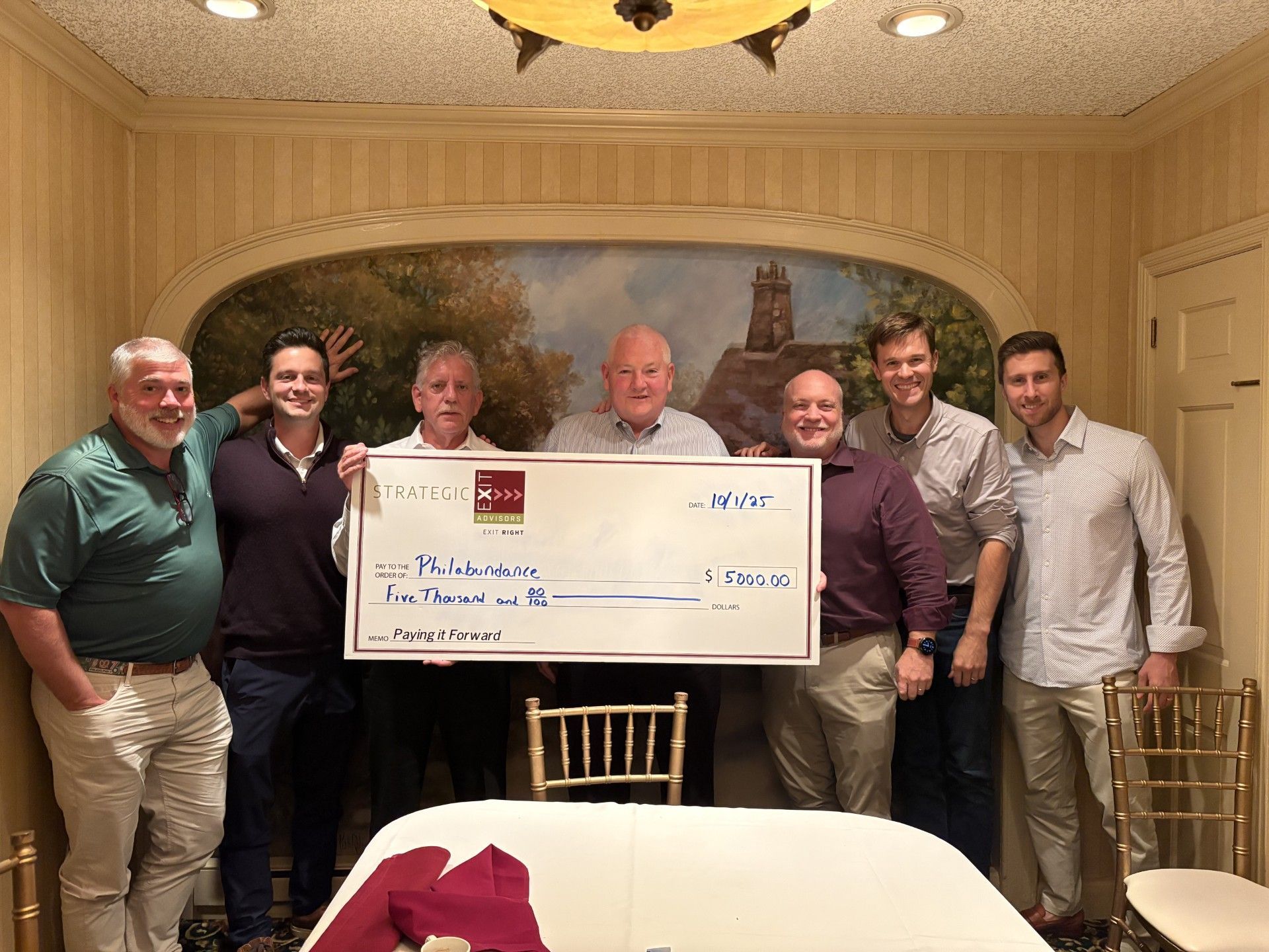 Group of men holding a large check indoors, smiling. They are standing near a painted wall, possibly a restaurant.