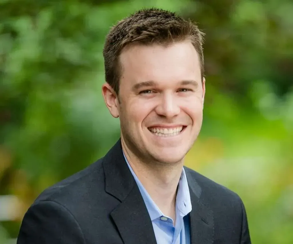 Man with short brown hair, smiling, wearing a blue shirt and black jacket. Outdoors, with blurred greenery.