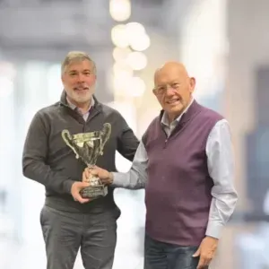 Two men holding a trophy. One man, gray sweater, holds the trophy. Older man, purple vest, smiles. Blurred indoor setting.