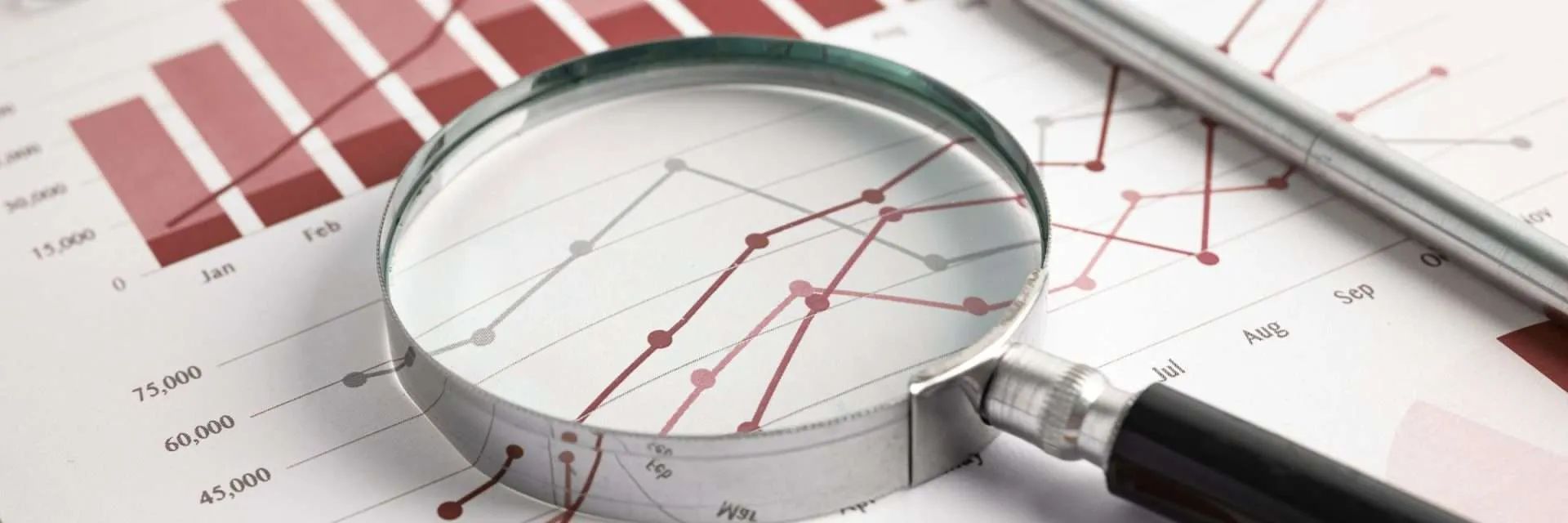 Magnifying glass over a financial graph with red lines and bar graph. A pen lies to the right.