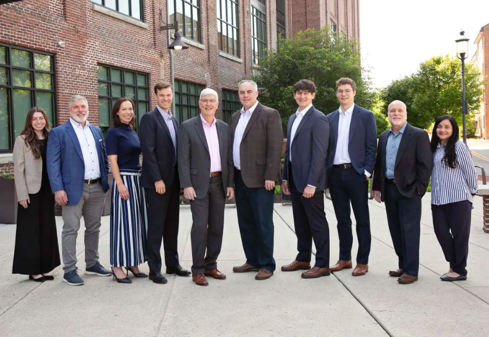 Group of people, business attire, pose outdoors in front of a brick building.